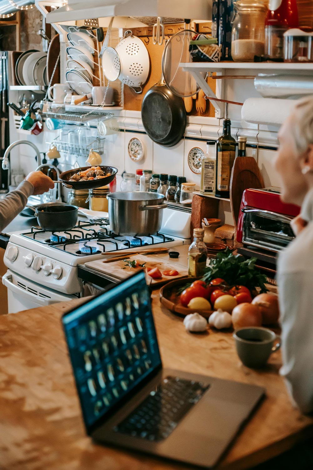 Woman using netbook in kitchen near husband cooking dishes on stove. Photo by Gary Barnes on Pexels.